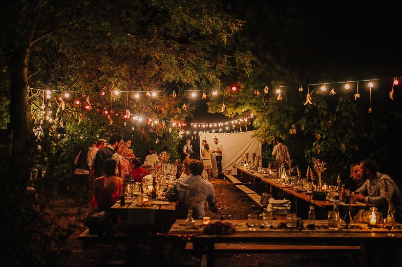 String lights illuminate an outdoor gathering with picnic tables and people enjoying a nighttime celebration.
