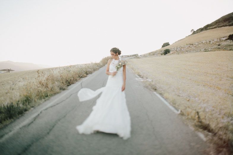 A flowing white wedding dress billows in the wind on a rural road surrounded by golden hills.