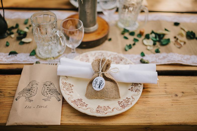 Rustic wedding table setting with kraft paper bags, floral plates, burlap favor bags and scattered greenery on wooden table.