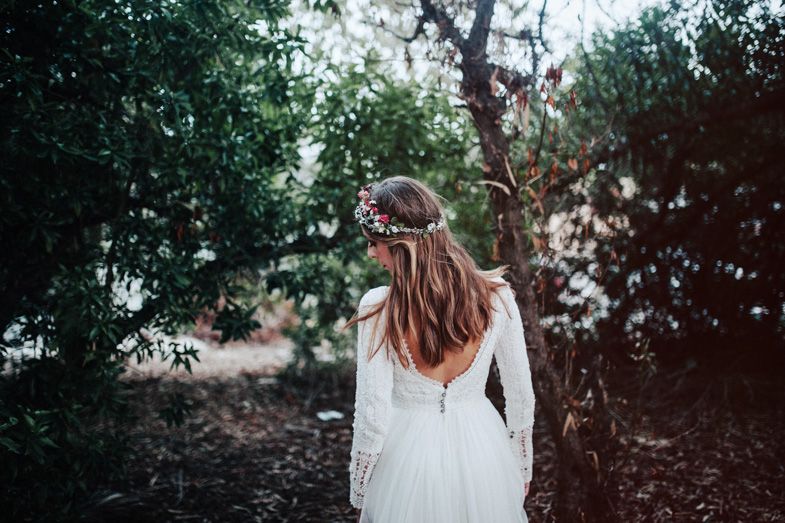 A bride in a white lace dress stands amid green foliage wearing a delicate floral crown.