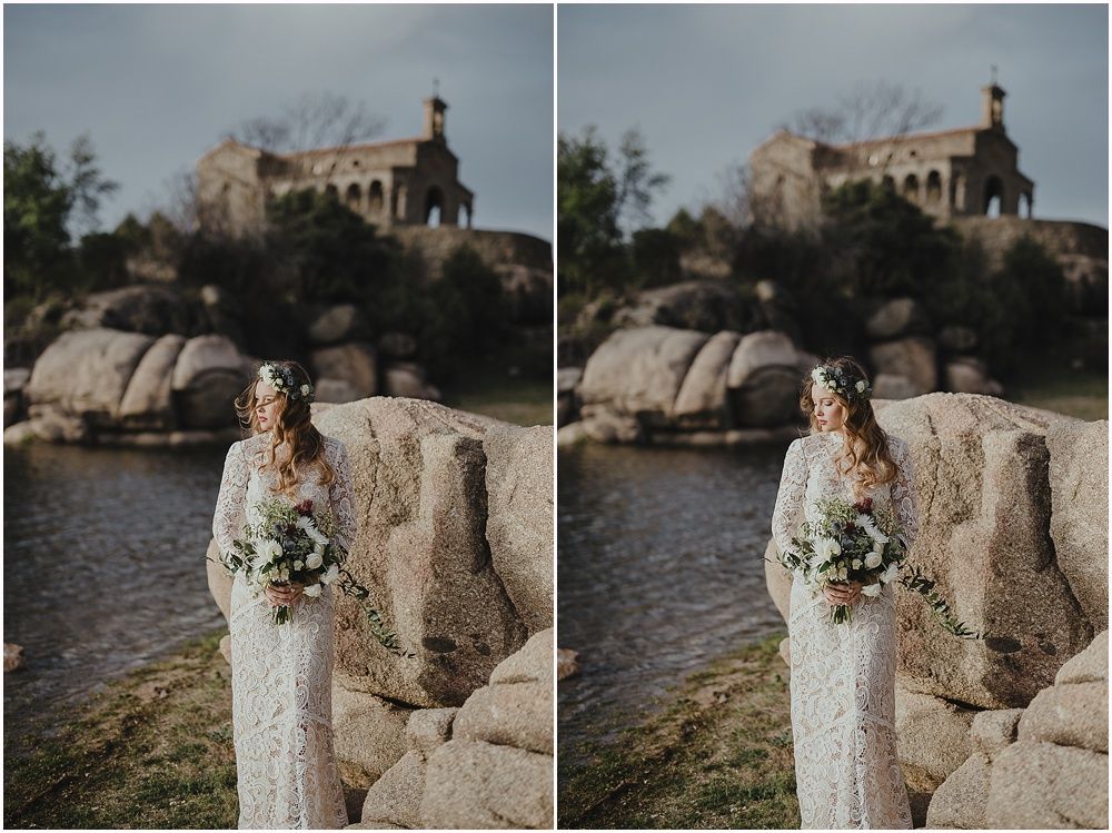 Bride in lace gown poses near stone church overlooking rocky landscape.