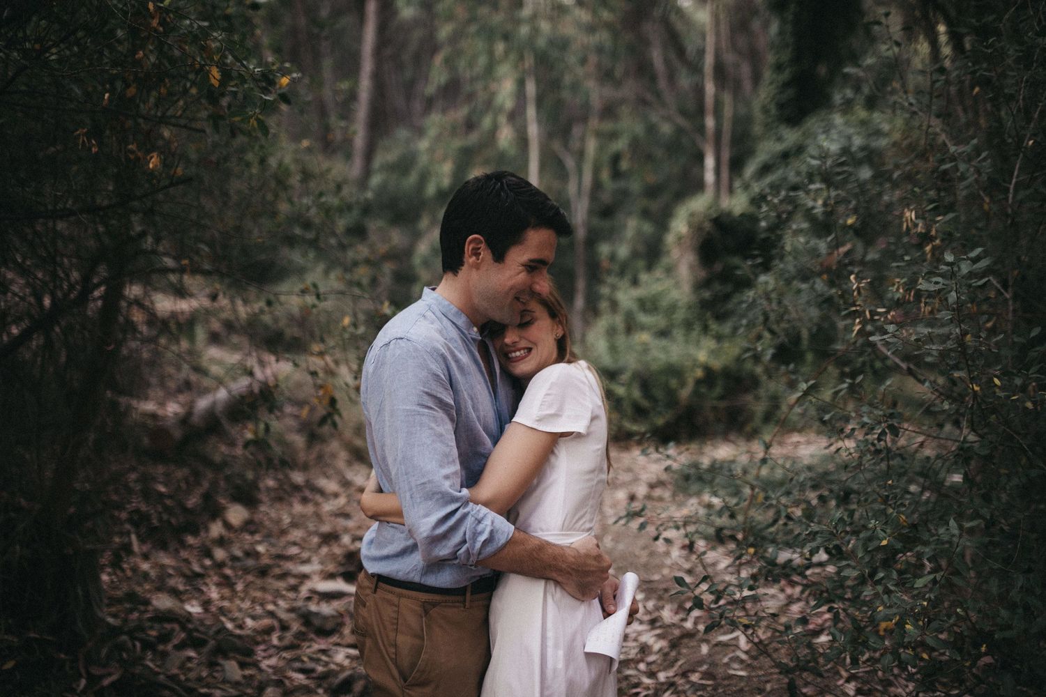 Couple embracing tenderly on a forest path wearing casual attire during an outdoor photoshoot.