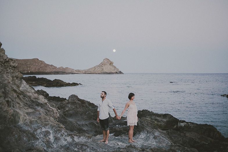 Two people stand on rocky coastline with ocean and mountains in background.