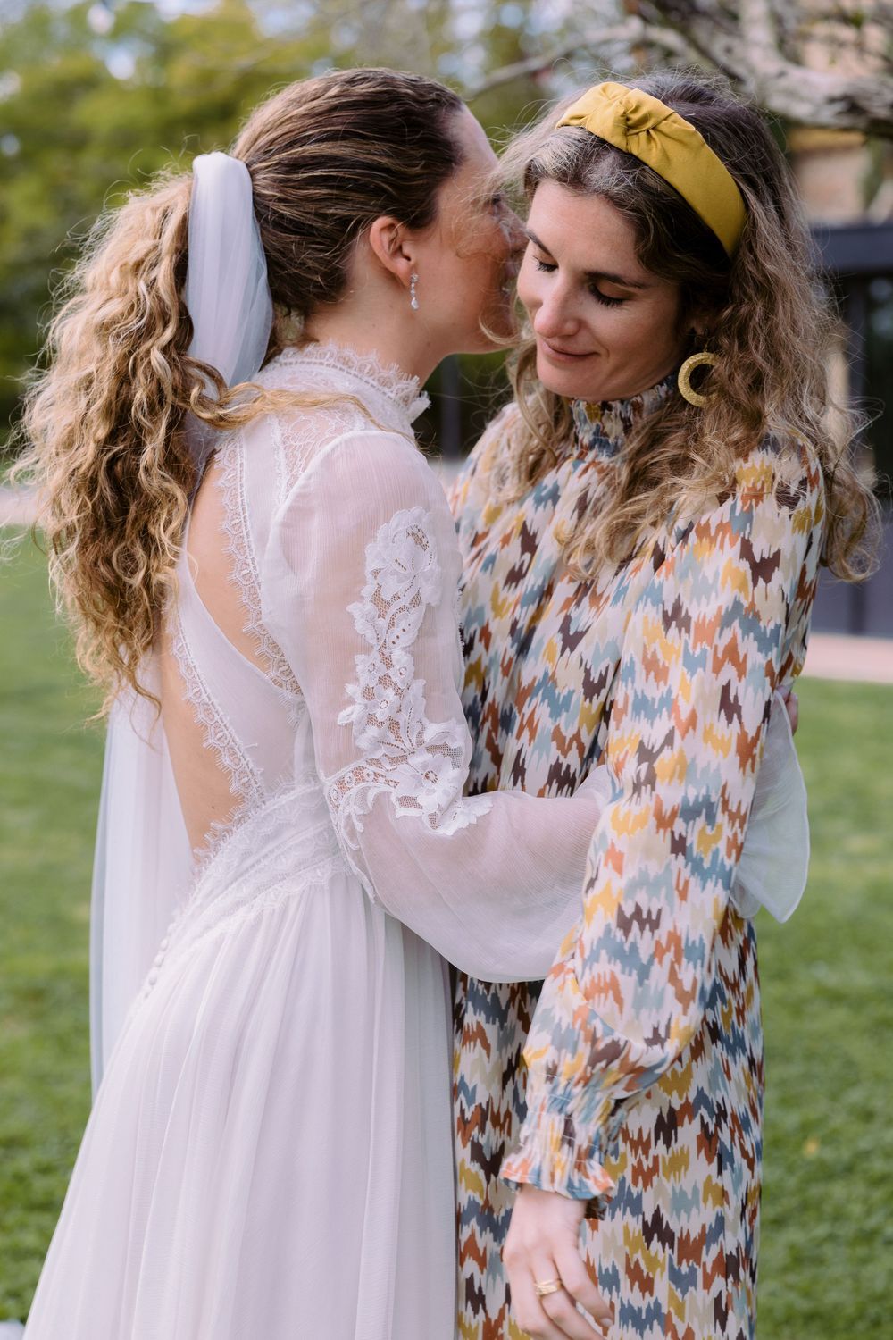 Two people share an intimate moment in a garden setting wearing white lace and floral attire.