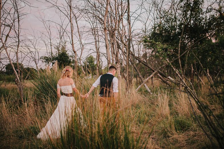 A couple holds hands while walking through tall grass at sunset.