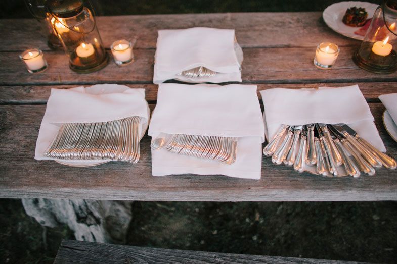 Folded cutlery wrapped in white napkins on a rustic wooden table with glowing candles.
