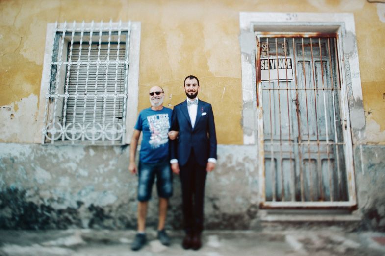 Two people pose against rustic wall with barred window and old door.