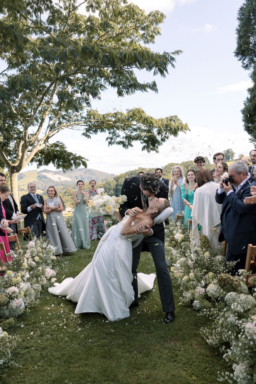 Wedding ceremony guests gather outdoors on a grassy hillside surrounded by white floral arrangements and trees.