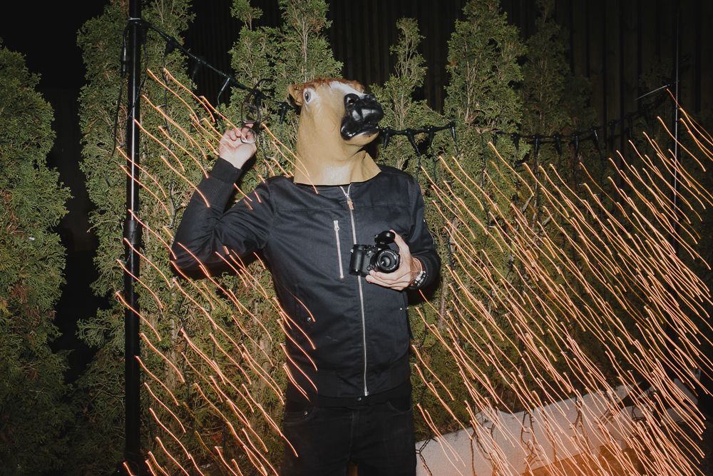Person in dark clothing wearing a horse mask poses dramatically against a lit bamboo backdrop.