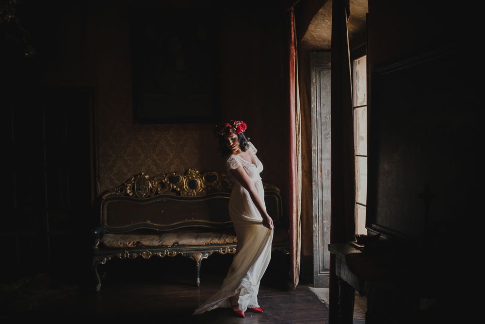 Figure in white dress poses gracefully by window in moody vintage room setting.