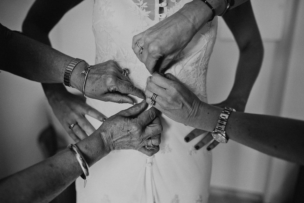Multiple hands working together to secure the back of a white wedding dress.