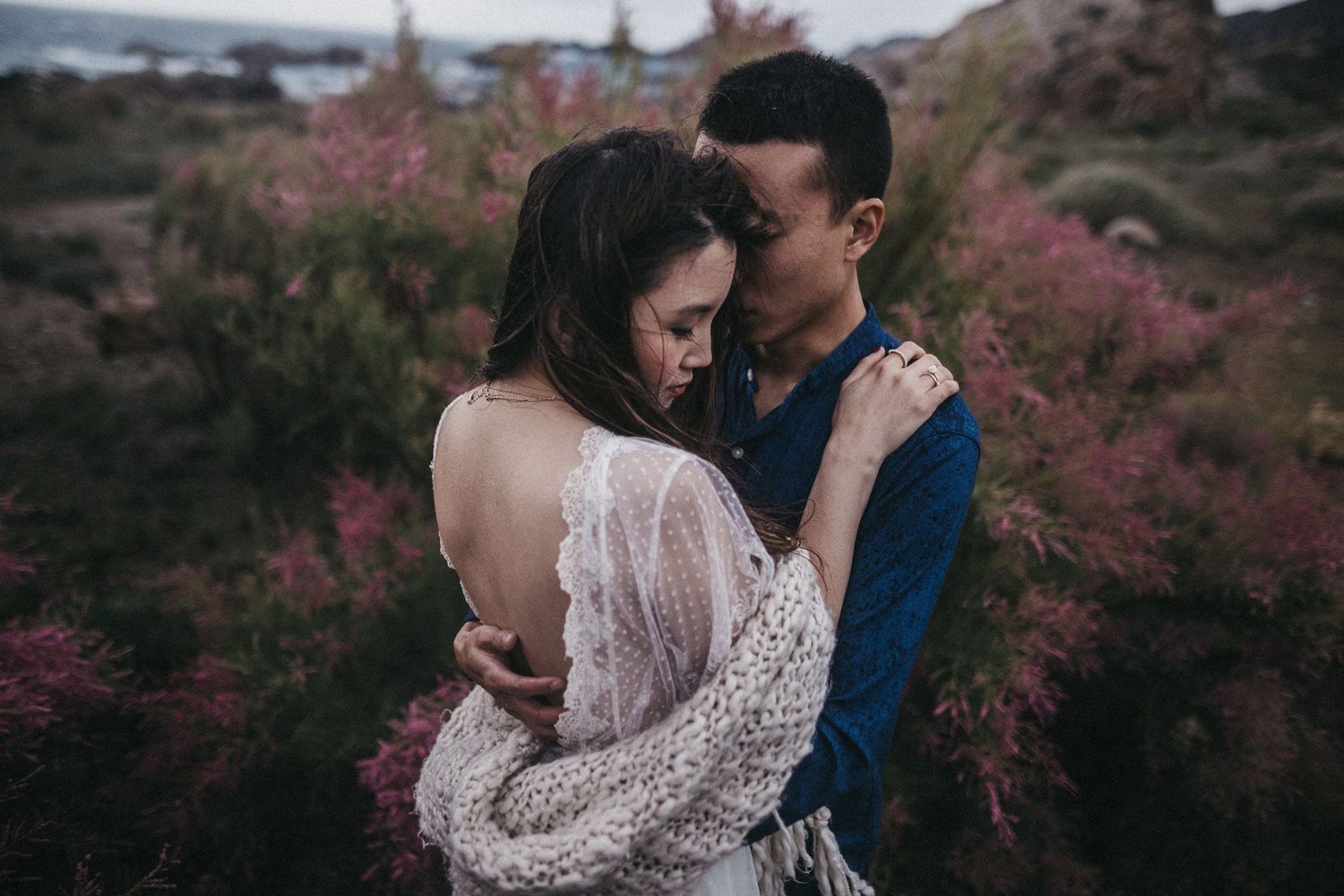 Romantic embrace captured in moody lighting with pink flowers in background.
