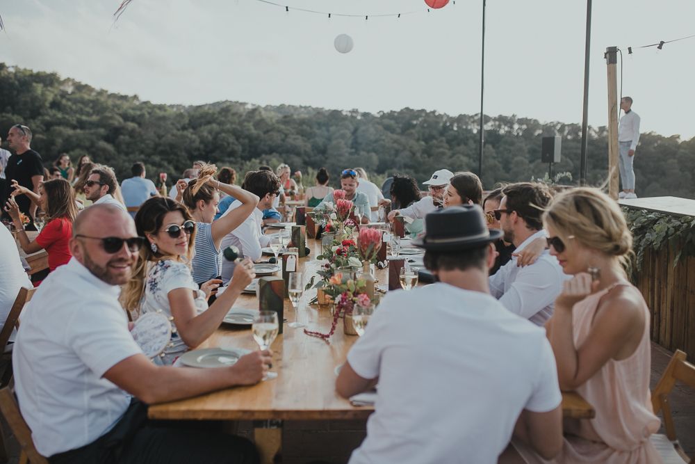 Guests enjoy an outdoor dining celebration at a long table on a sunny day with hills in the background.