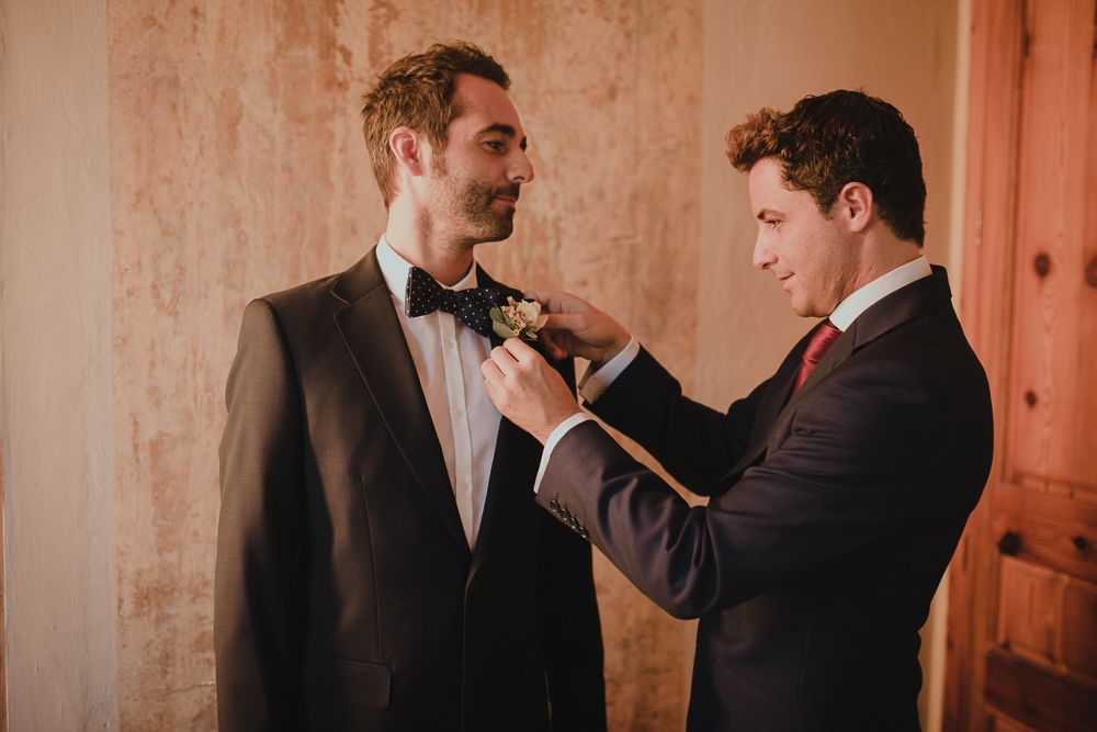 Wedding preparation scene showing bow tie adjustment against a warm-toned wall.