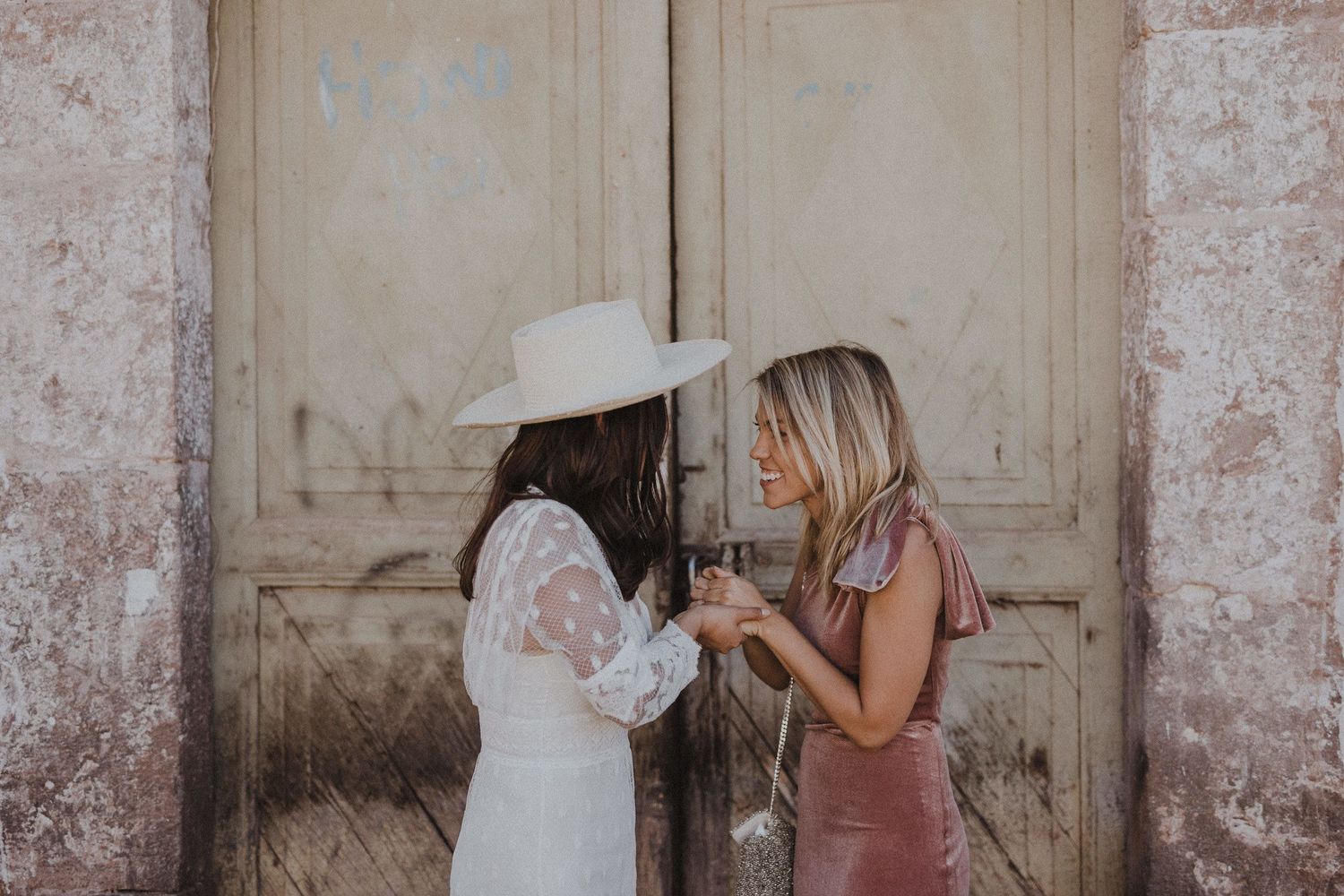 Two guests in white and blush dresses share a laugh against an antique door.