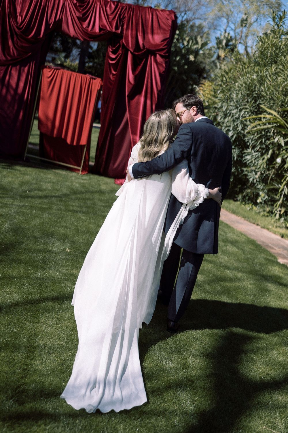 A couple embraces during an outdoor wedding ceremony with red curtains and greenery in the background.