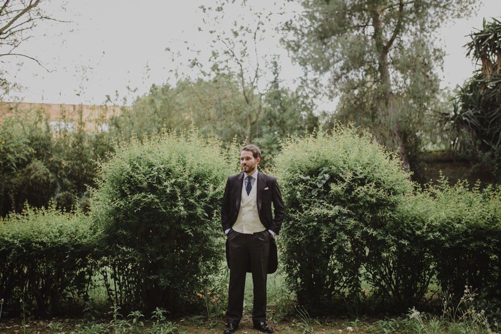 A groom in a dark suit stands against green garden hedges on a wedding day.