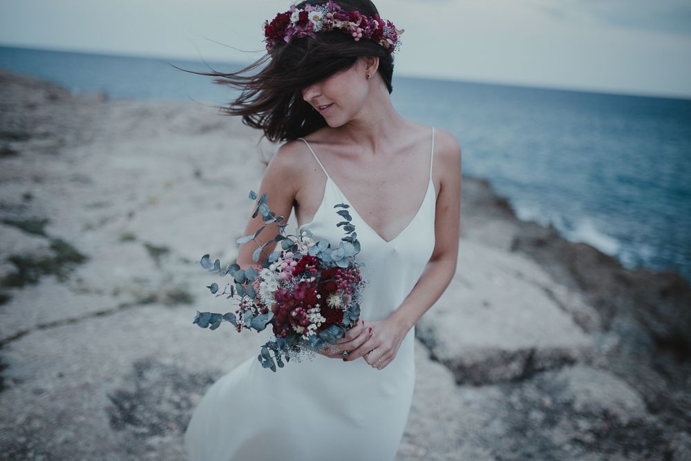 A flowy white dress and burgundy floral crown complement a seaside bridal portrait.