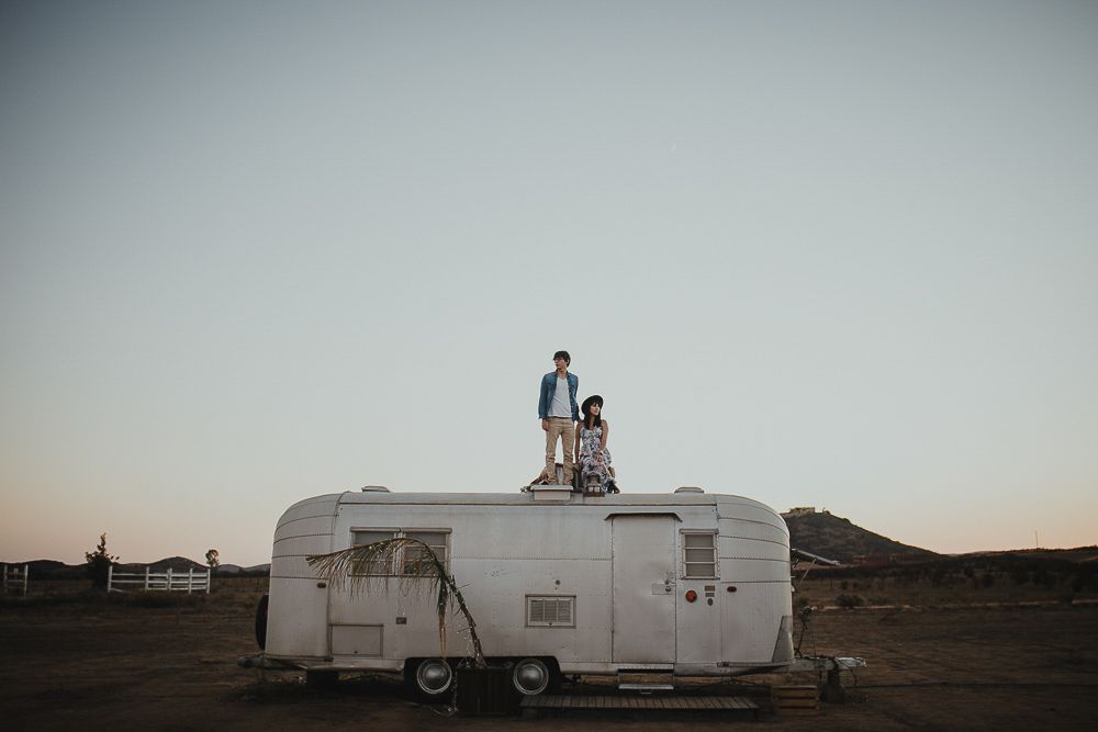 A couple stands on top of a vintage Airstream trailer in a desert setting at dusk.