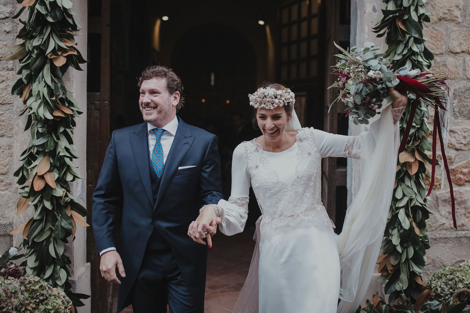Wedding couple emerges joyfully through doorway decorated with greenery garlands.