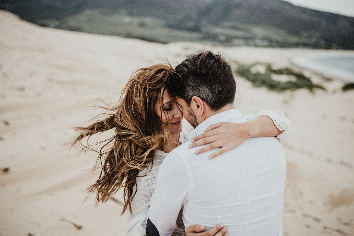 A romantic couple embraces on a windswept beach with mountains in the background.