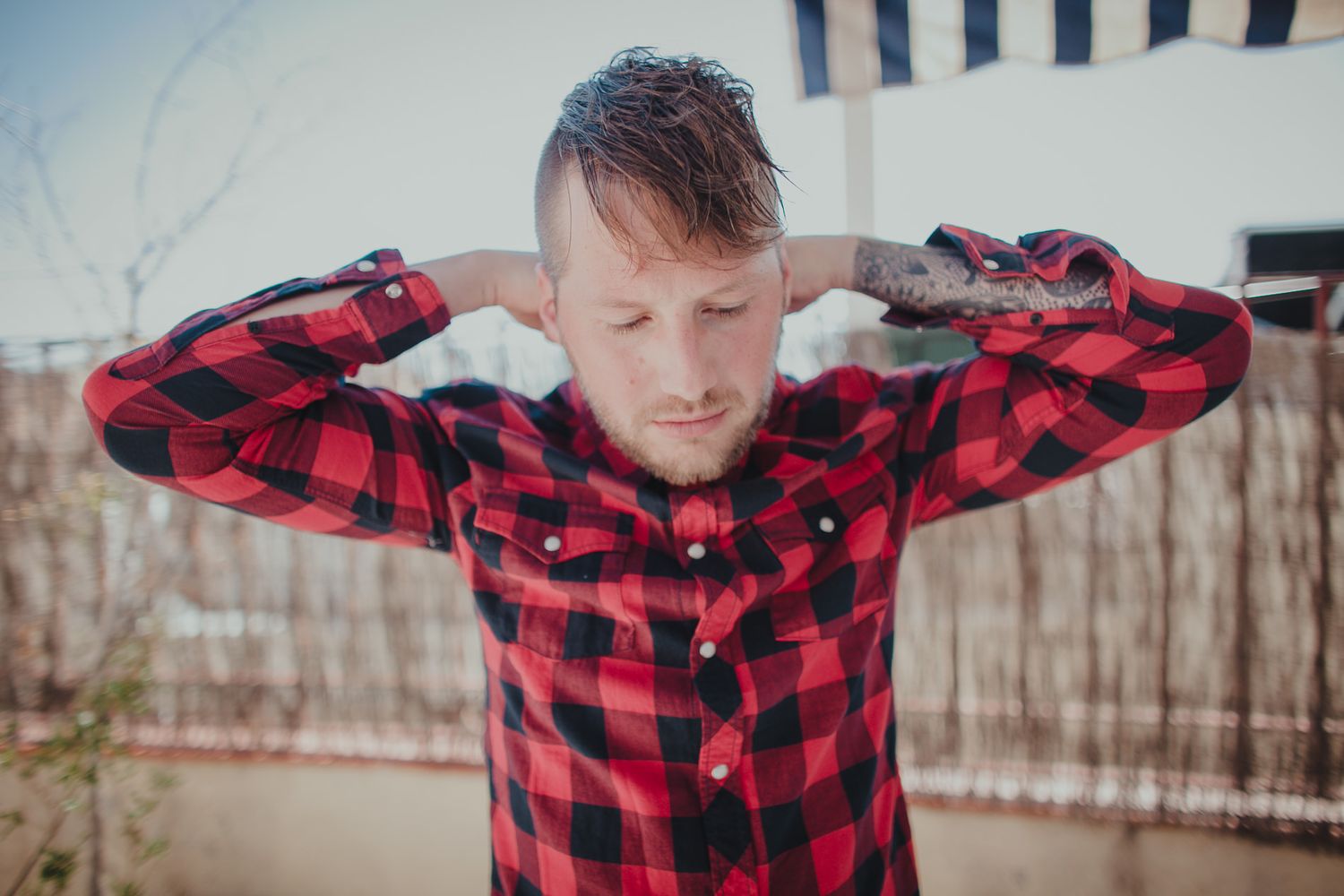 Person in red plaid flannel shirt standing outdoors with hands behind head.