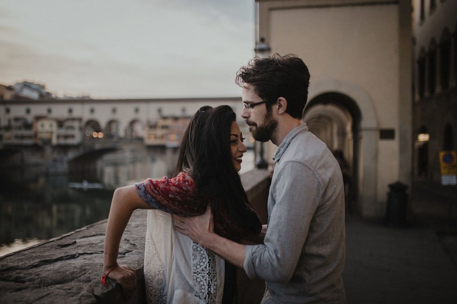 Romantic couple embracing near historic bridge in Florence at sunset.