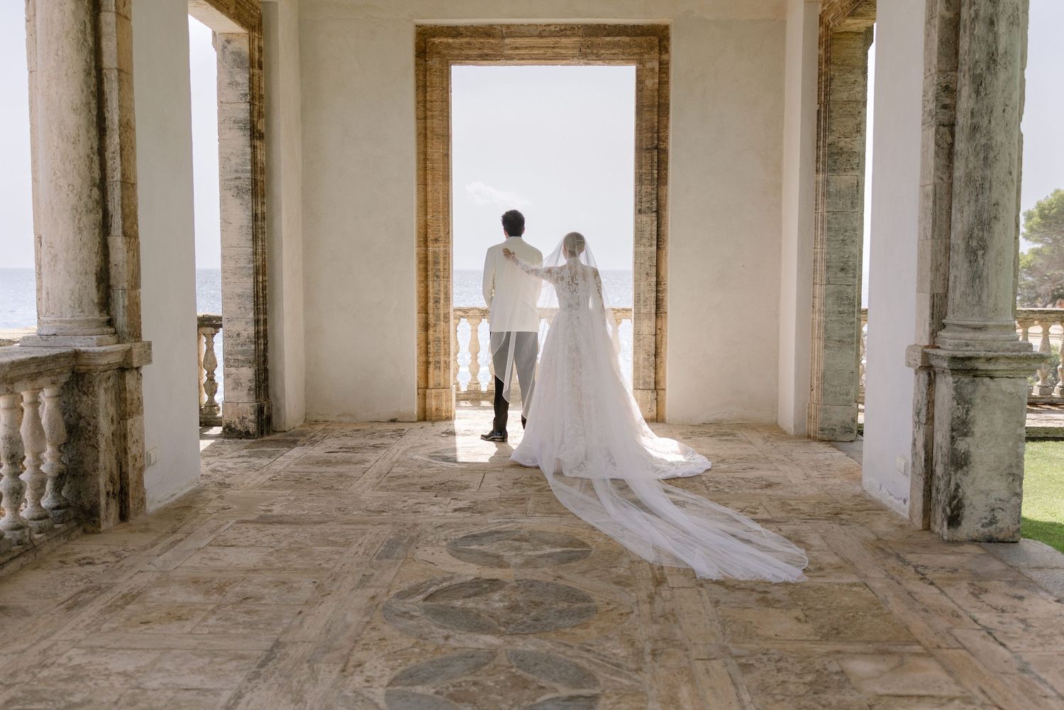 Bride and groom stand in elegant stone archway overlooking ocean at luxury wedding venue.