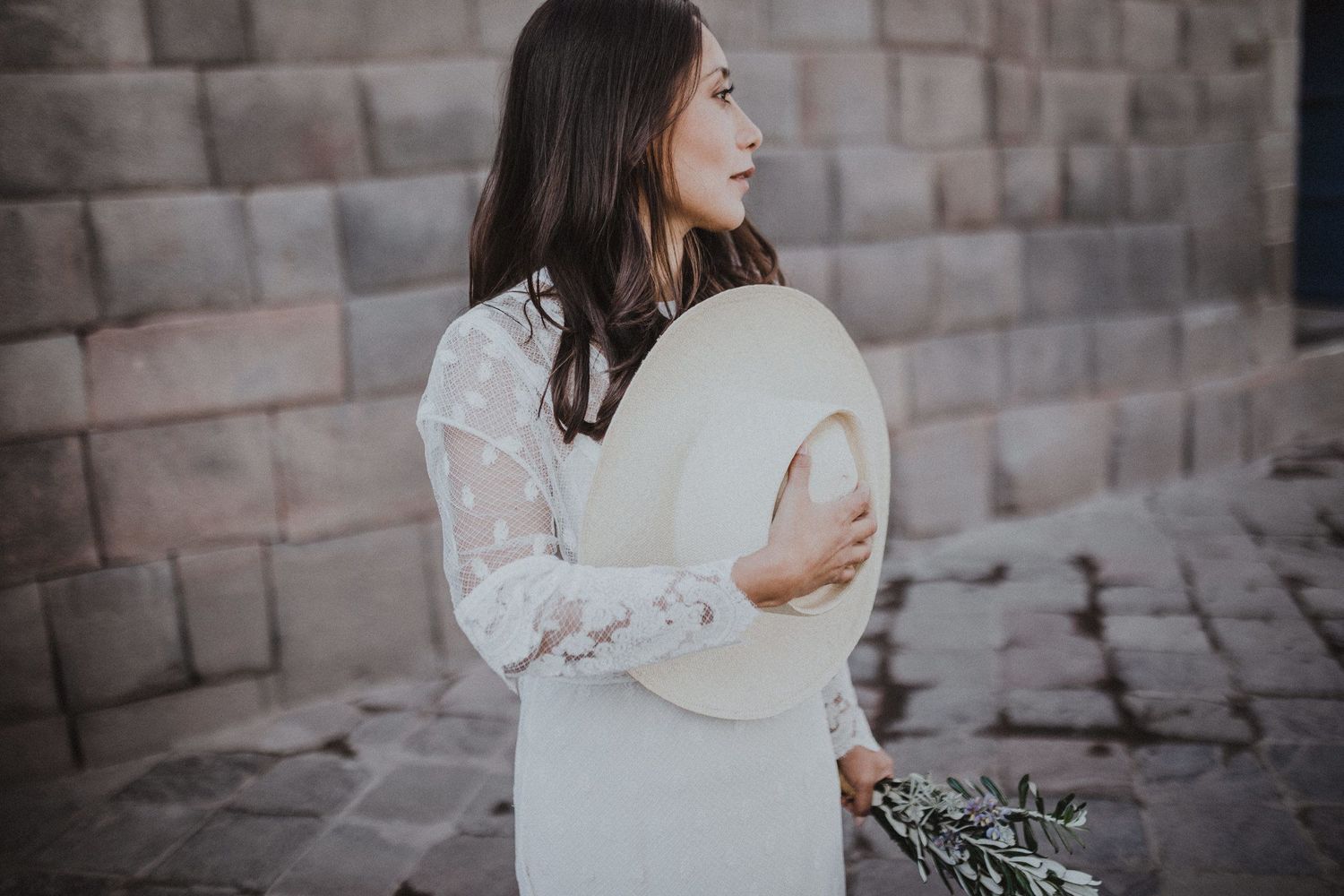 Woman in white lace dress holds a light-colored hat while standing against stone wall.
