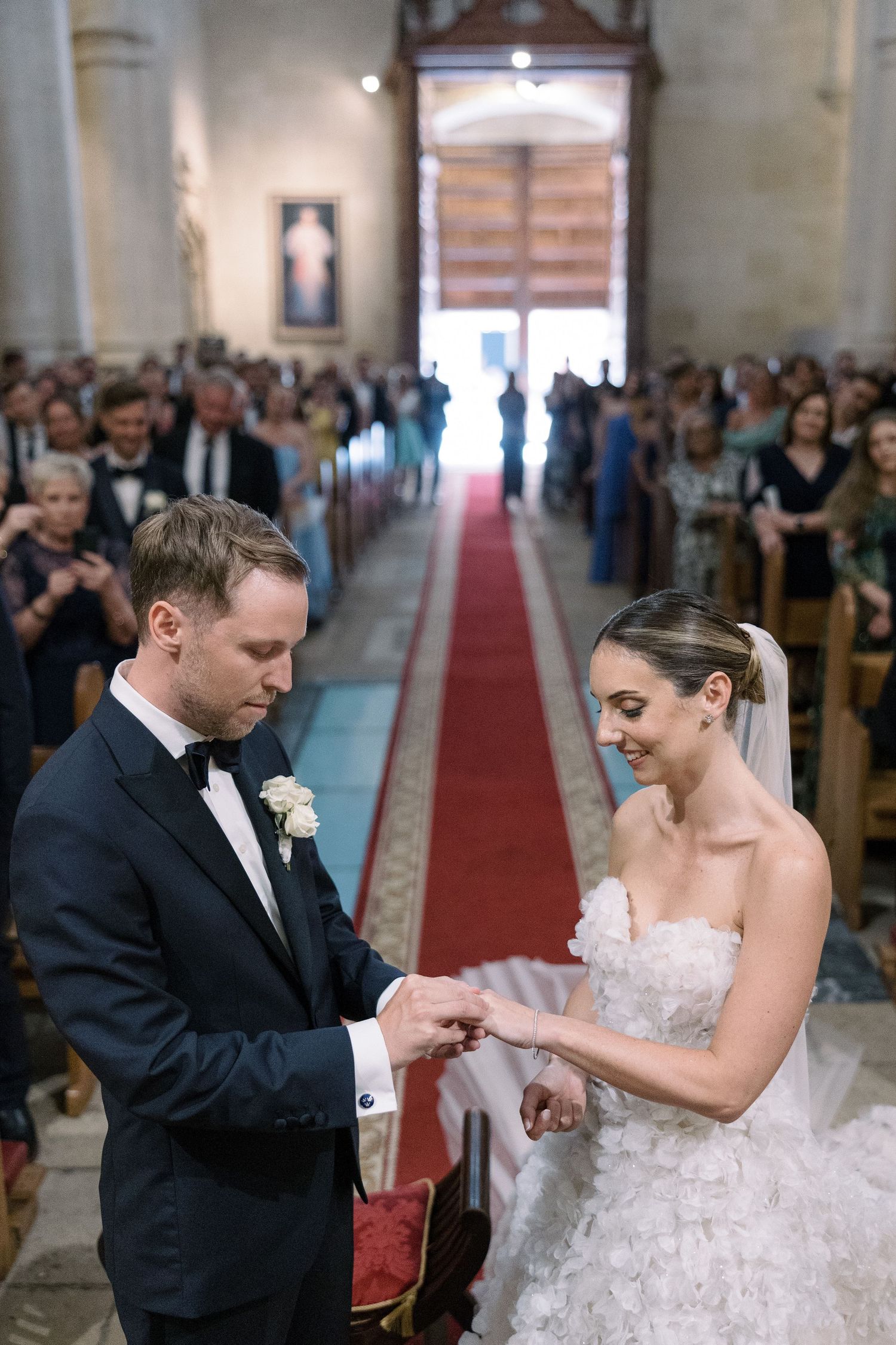 A couple exchanges rings during their wedding ceremony in a grand church with guests looking on.