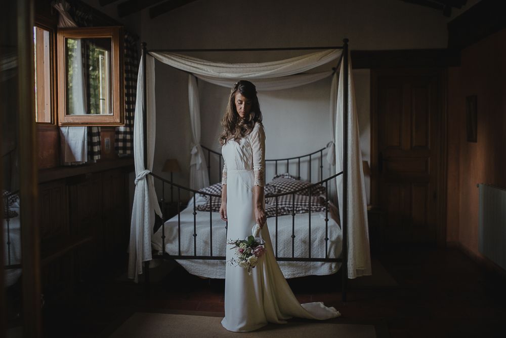 Elegant bride in long white dress stands before draped bed canopy in rustic wooden room setting.