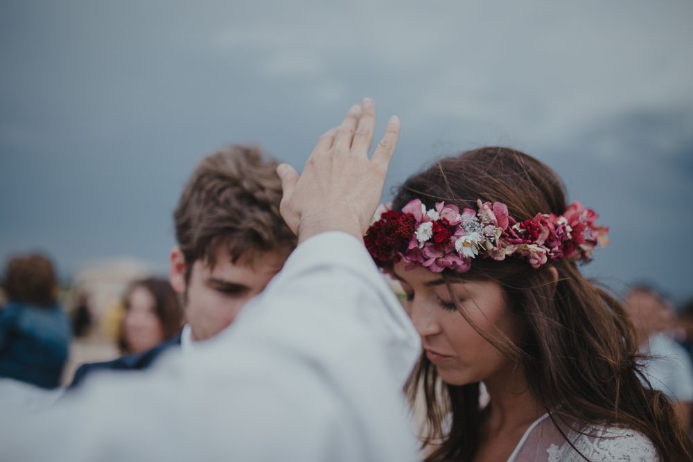 A blessing is given during an outdoor wedding ceremony as a bride wearing a pink floral crown bows her head.