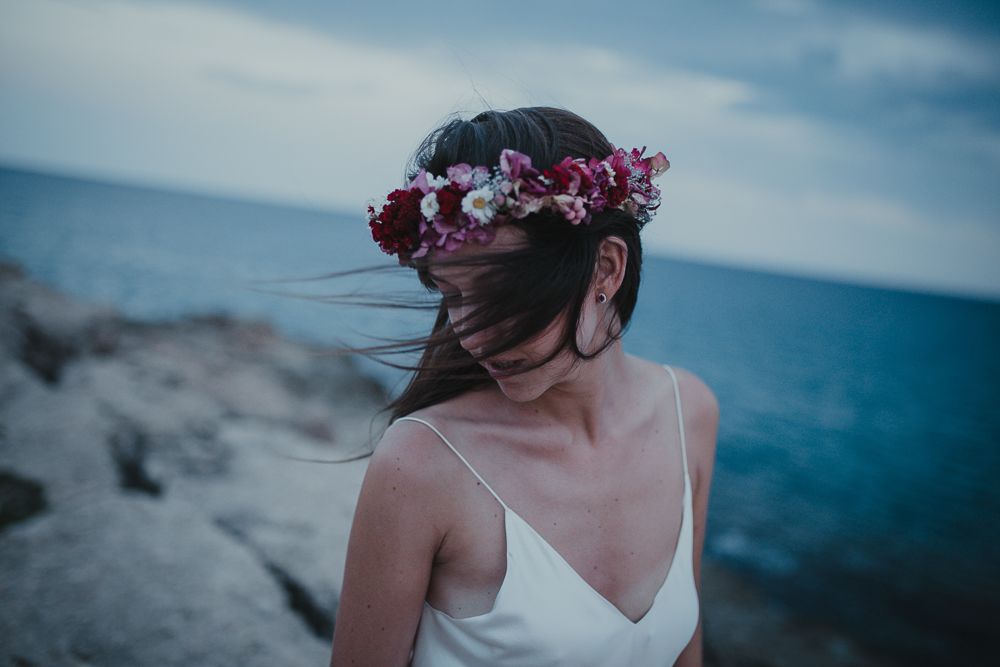 A bride in a white dress wears a burgundy floral crown against an ocean backdrop at sunset.