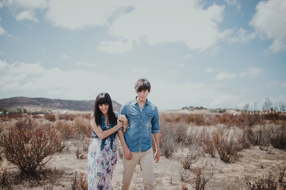 Two people in coordinating blue and floral outfits walk through desert brush together.