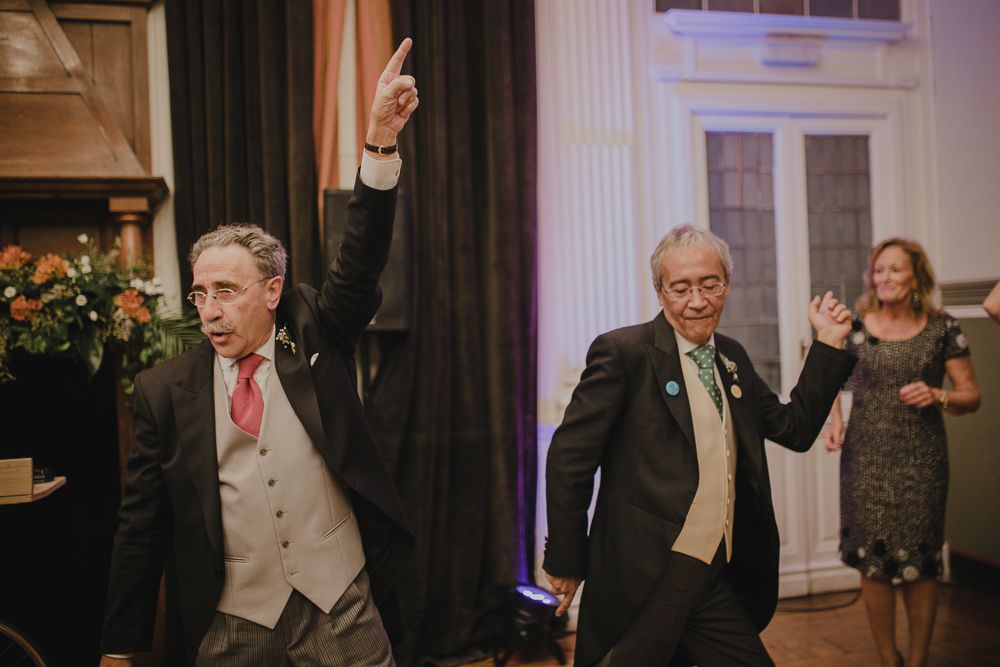 Two men in formal attire raise their hands in celebration at an indoor event.