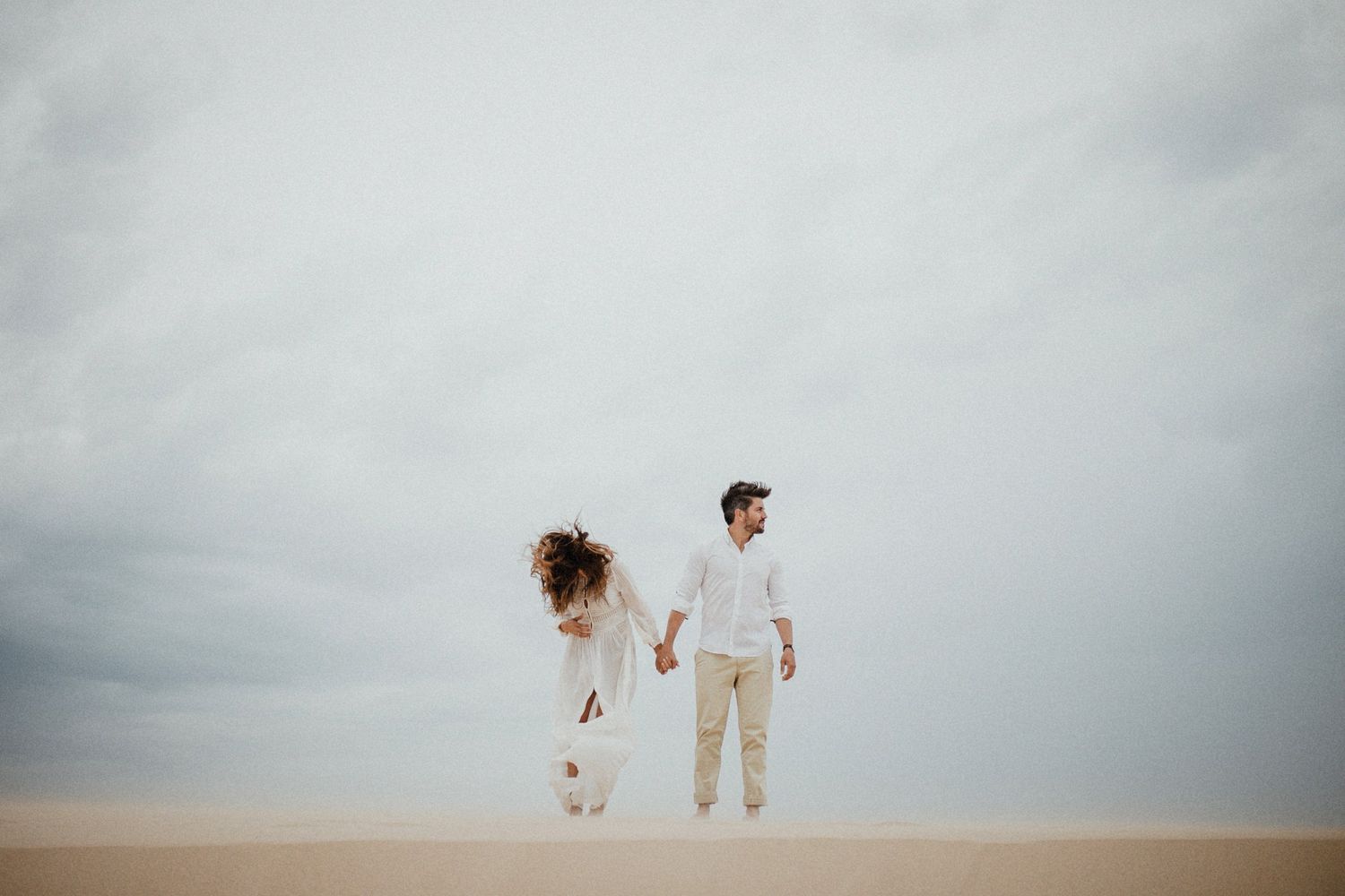 Couple holding hands while walking on sandy dunes with overcast sky in background.