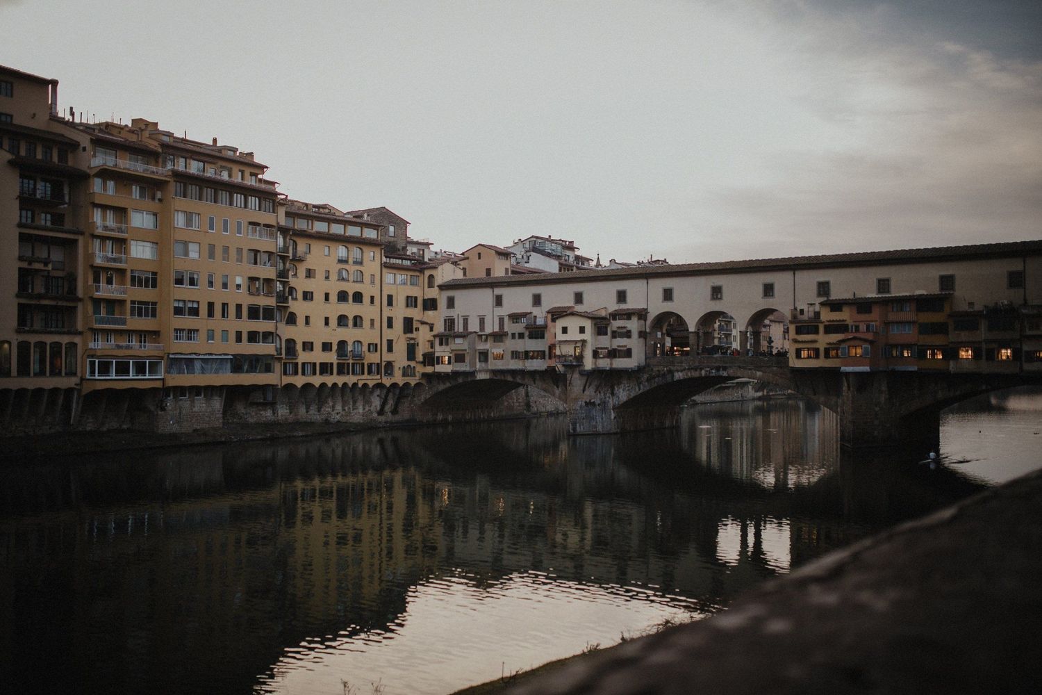Historic Ponte Vecchio bridge spans the Arno River at dusk in Florence, Italy with reflections in calm water.