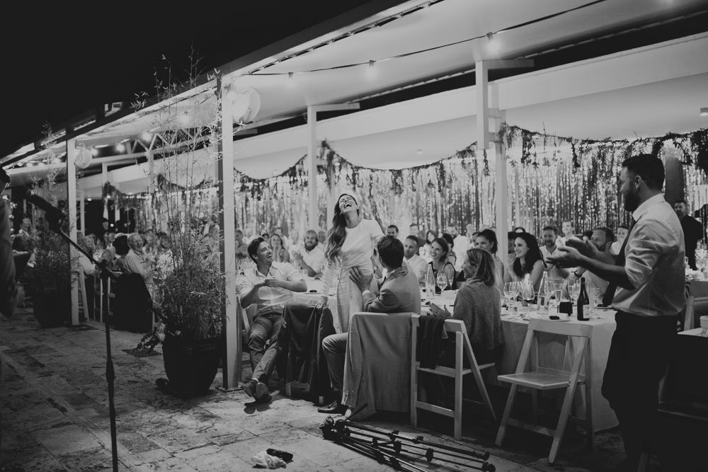 Wedding reception guests dine under a decorated outdoor tent with string lights and streamers.