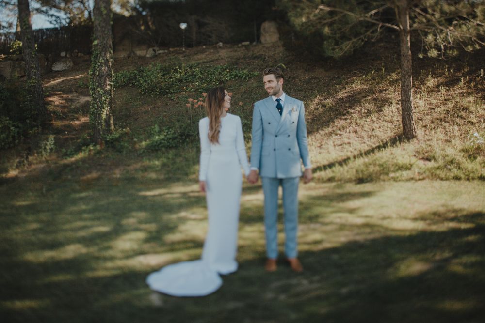 Wedding couple standing in sunlit forest clearing wearing blue suit and white dress.