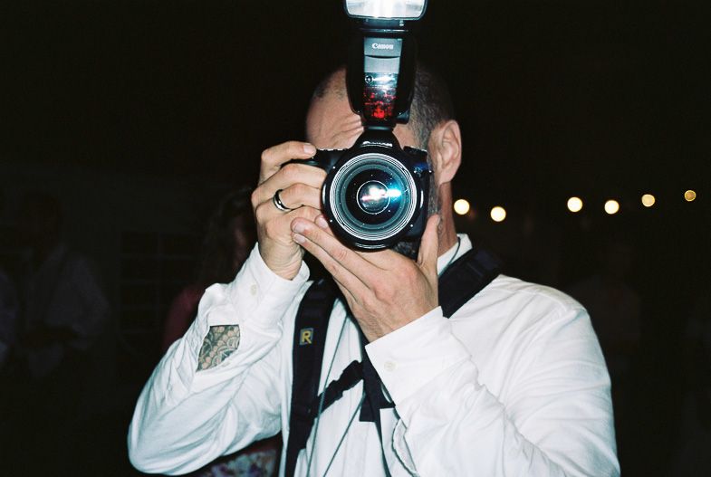 Photographer in white shirt holding camera with mounted flash photographing in low light conditions.