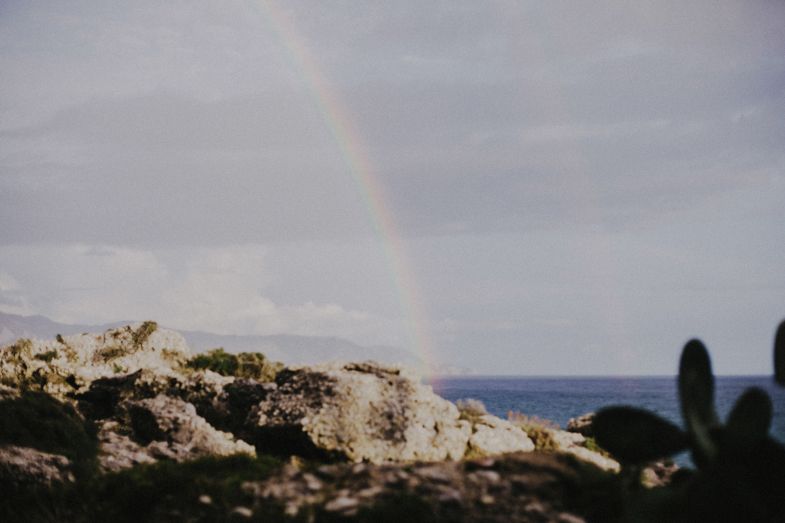 A rainbow arches over rocky coastal cliffs against a moody gray sky and ocean backdrop.