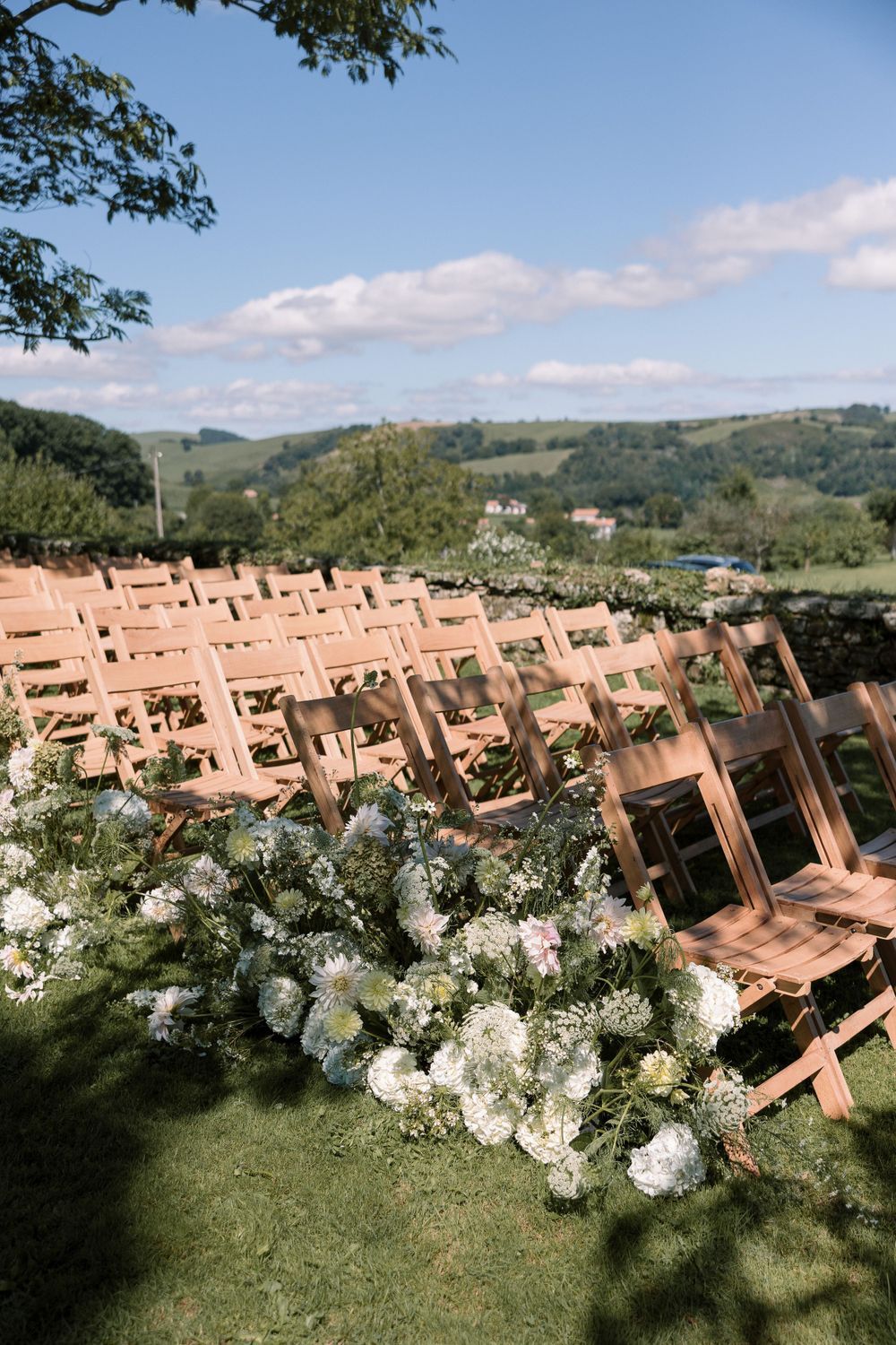 Rows of wooden chairs decorated with white floral arrangements set up for outdoor wedding ceremony.