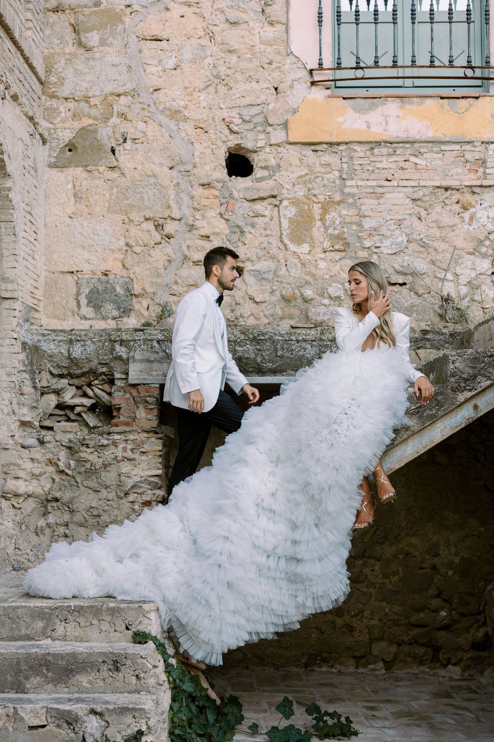 A dramatic wedding photo on stone steps with a flowing white ruffled dress against an aged wall.