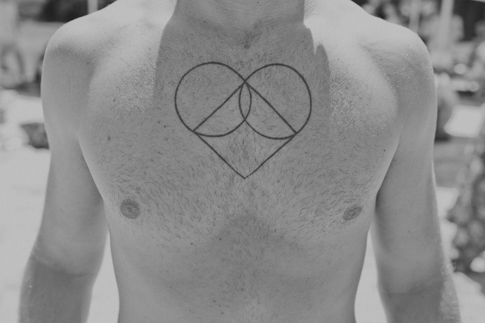Close-up black and white photo of a minimalist heart and infinity symbol tattoo on a person's chest.