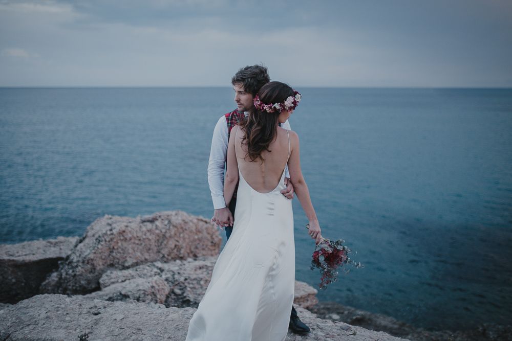 Couple stands on rocky cliff overlooking vast blue ocean in wedding attire.