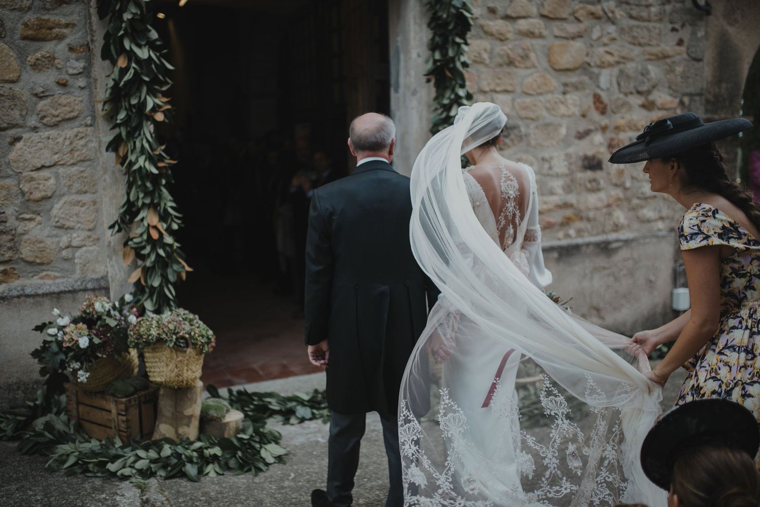 Long flowing wedding veil catches the breeze against stone church entrance decorated with florals.