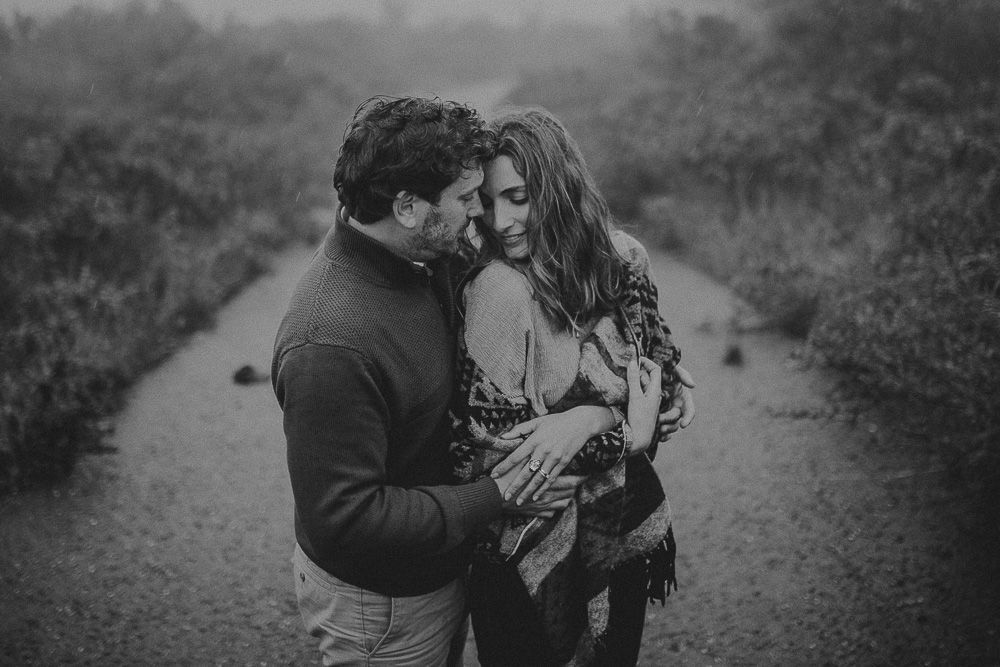 A tender black and white portrait of a couple embracing on a foggy countryside path.
