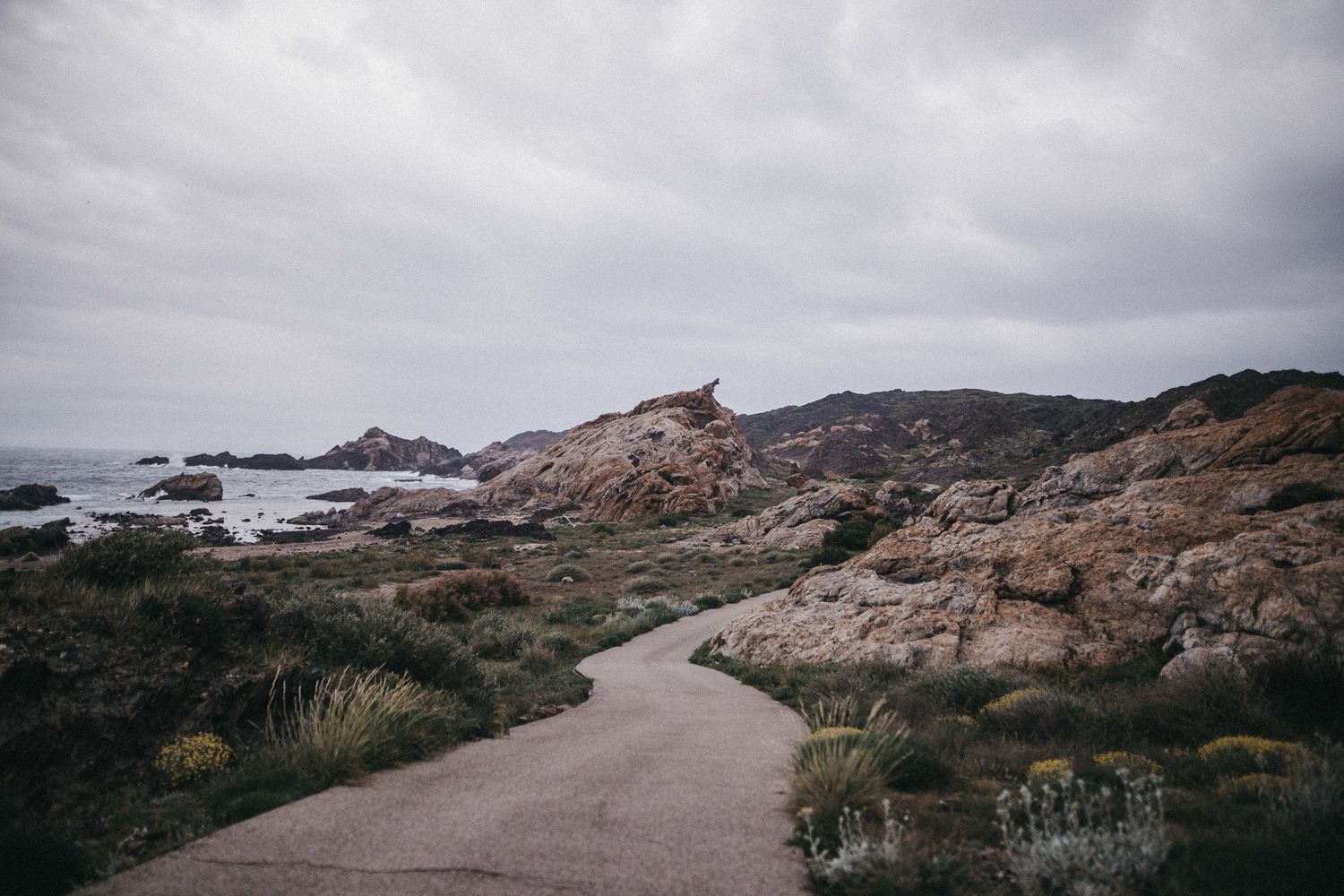 Winding coastal path through rocky terrain with ocean views and overcast sky.