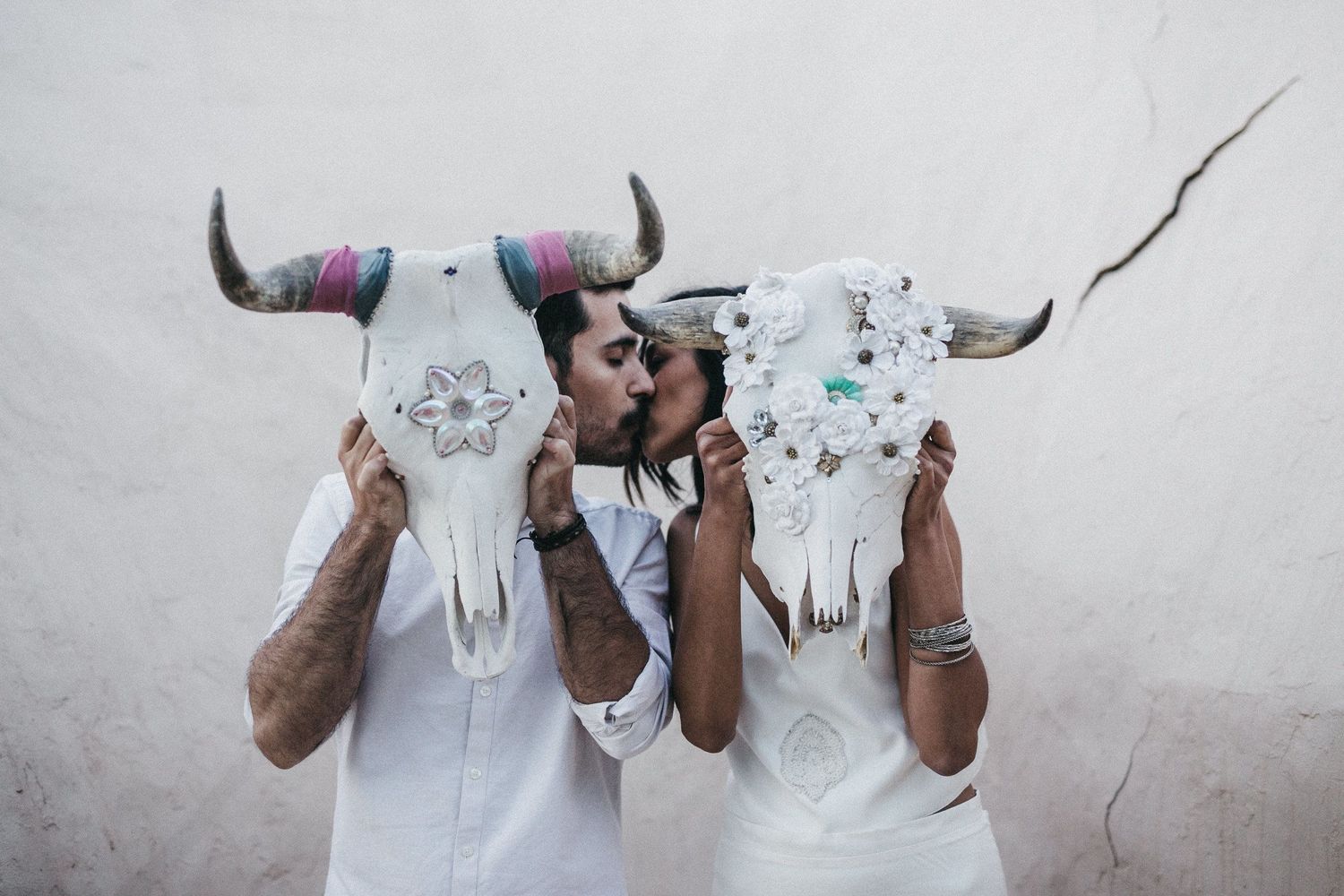 Two people holding decoratively painted animal skulls with horns against a white wall.