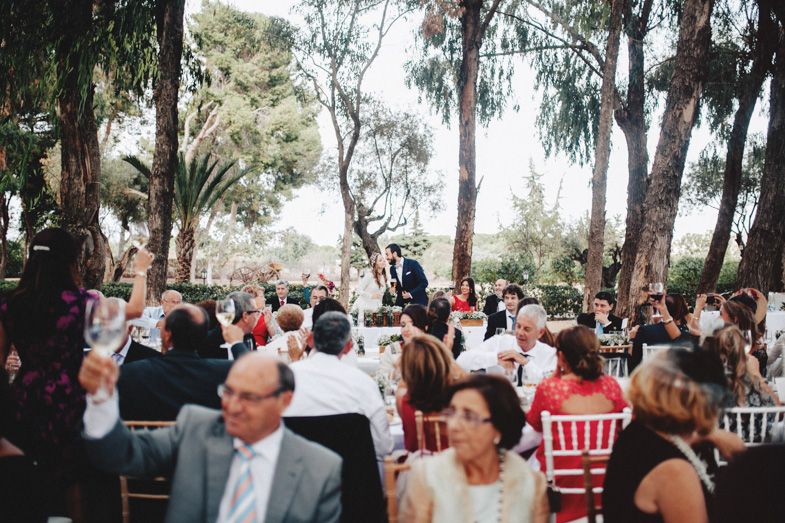 An outdoor wedding ceremony setup in a wooded area with guests seated on white chairs among tall trees.